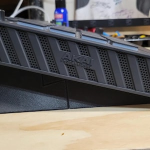 May include: A dark grey Akai Professional music production controller resting on a black angled stand. The device features a perforated side panel with diagonal slats, a top-mounted control knob, and a series of square pads. The brand name Akai Professional is embossed on the side of the unit.