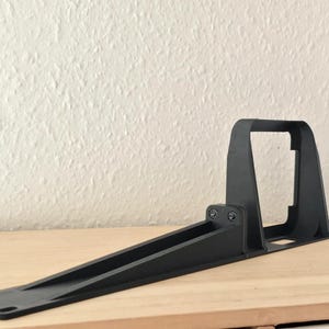 May include: A matte black plastic mounting bracket featuring a long, tapered base with pre-drilled holes at the ends and a vertical, rectangular support structure attached to the center with two visible screws for secure installation.