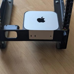 May include: A silver Apple Mac Mini computer mounted within a black metal server rack frame. The device features the iconic black Apple logo on its top surface and is positioned securely between the vertical rails of the rack mount system.
