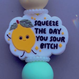 May include: A silicone beaded keyring featuring a white cloud-shaped charm with a yellow lemon character and the black text SQUEEZE THE DAY YOU SOUR BITCH. The charm is accented by small yellow stars and is strung between round yellow and teal beads.