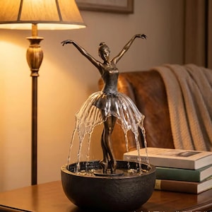 Könnte beinhalten: Ein bronzefarbener Ballerina-Zimmerbrunnen, der eine Tänzerin mit erhobenen Armen zeigt. Wasser fließt aus dem Taillenbereich und erzeugt einen rockartigen Effekt, der in ein dunkles, kreisförmiges Becken kaskadiert. Die Figur steht auf einem Sockel.