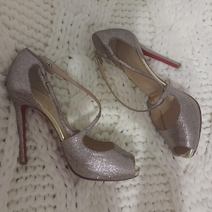 May include: A pair of silver glitter peep-toe high heels with thin ankle straps and a distinctive red sole. The shoes feature a cut-out design on the sides, a slim stiletto heel, and a metallic gold-toned accent at the open toe area.