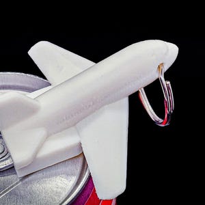 May include: A white airplane-shaped bottle opener made of a solid material, featuring a sleek fuselage and wings, with a silver metal ring attached to the nose section, positioned on top of a metallic beverage can lid against a black background.