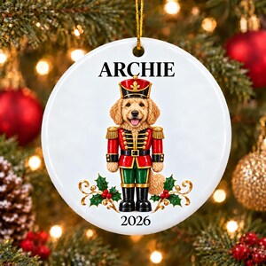 Może przedstawiać: Okrągła biała ceramiczna ozdoba świąteczna przedstawiająca psa rasy golden doodle przebranego za dziadka do orzechów w czerwono-zielonym mundurze. Imię ARCHIE jest nadrukowane czarną czcionką nad psem, a rok 2026 pod psem.