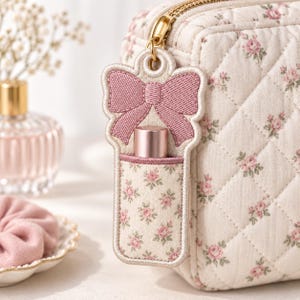 May include: A quilted cream-coloured cosmetic bag with a small pink floral pattern. Attached to the gold zip pull is a matching fabric holder featuring an embroidered pink bow at the top and a pocket containing a small metallic pink lipstick tube.
