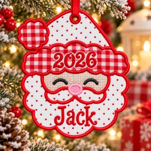 May include: A white and red embroidered Christmas ornament shaped like a Santa face. The design features a red and white gingham hat brim with the year 2026, a pink nose, closed eyes, and the name Jack embroidered in red script below the beard.