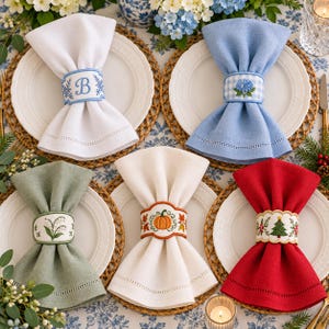May include: Five linen napkins folded and secured with embroidered napkin rings. Designs include a blue letter B, blue hydrangea, lily of the valley, an orange pumpkin with autumn leaves, and a green Christmas tree with red berries.