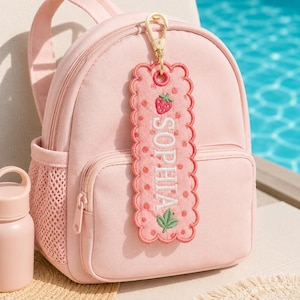 May include: A light pink backpack with a matching pink embroidered luggage tag attached by a gold lobster clasp. The rectangular tag features a scalloped edge, a strawberry motif, and the name SOPHIA stitched in white capital letters.