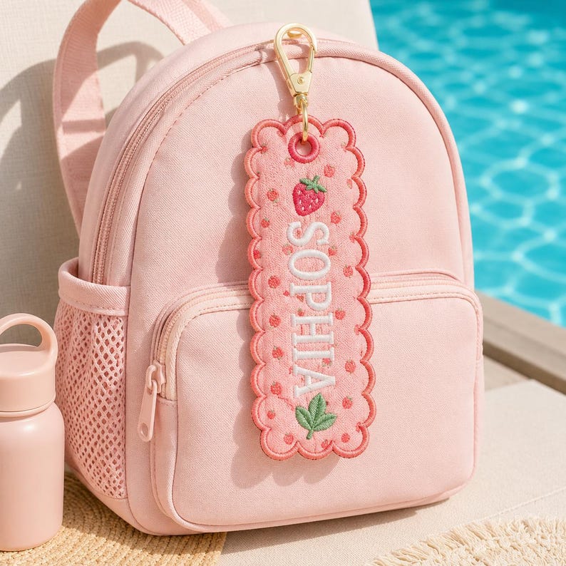 May include: A light pink backpack with a matching pink embroidered luggage tag attached by a gold lobster clasp. The rectangular tag features a scalloped edge, a strawberry motif, and the name SOPHIA stitched in white capital letters.