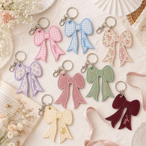 May include: A collection of eight embroidered bow keychains in various colors and patterns, including gingham, floral, and solid fabrics. Each bow features a metal lobster clasp and is personalized with a name embroidered on one of the tails.