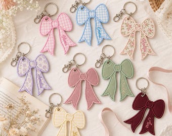 May include: A collection of eight embroidered bow keychains in various colors and patterns, including gingham, floral, and solid fabrics. Each bow features a metal lobster clasp and is personalized with a name embroidered on one of the tails.