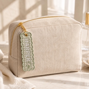 May include: A beige rectangular cosmetic bag made of a textured fabric with a gold zipper. Attached to the zipper pull is a sage green scalloped fabric tag featuring the name HARPER embroidered in white, along with a small floral design.
