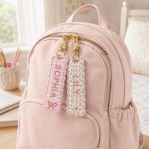 May include: A light pink backpack featuring two rectangular embroidered bag tags attached to the gold zipper pulls. One tag has a pink gingham pattern with the name SOPHIA and a bow, while the other has a floral print with the name LILY and a daisy.