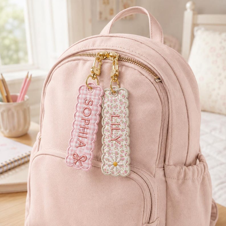 May include: A light pink backpack featuring two rectangular embroidered bag tags attached to the gold zipper pulls. One tag has a pink gingham pattern with the name SOPHIA and a bow, while the other has a floral print with the name LILY and a daisy.