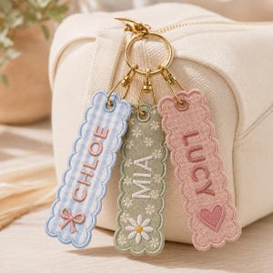 May include: Three rectangular fabric keychains with scalloped edges attached to a gold-toned metal ring. The left keychain is light blue gingham with the name CHLOE and a bow. The center is sage green with daisies and the name MIA. The right is pink with the name LUCY and a heart.