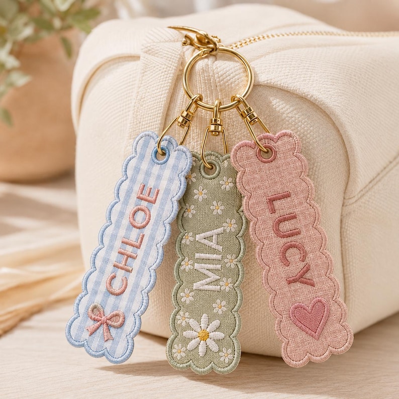 May include: Three rectangular fabric keychains with scalloped edges attached to a gold-toned metal ring. The left keychain is light blue gingham with the name CHLOE and a bow. The center is sage green with daisies and the name MIA. The right is pink with the name LUCY and a heart.