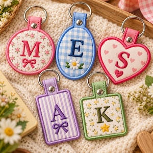May include: Five embroidered keychains with metal rings. One round with pink floral print and letter M, one oval with blue gingham and letter E, one heart with pink hearts and letter S, one rectangular with purple stripes and letter A, and one square with yellow floral print and letter K.