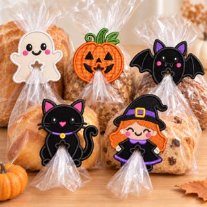 May include: Five embroidered Halloween-themed bag clips featuring a white ghost, an orange pumpkin, a black bat, a black cat, and a witch with orange hair. Each clip is attached to a clear plastic bag containing a loaf of bread or baked goods.