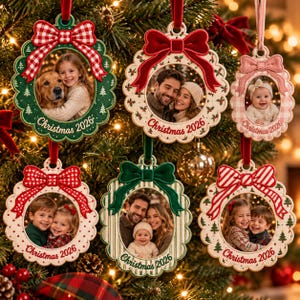 May include: Six oval-shaped fabric Christmas ornaments featuring photo inserts of families and pets. Each ornament has a decorative bow at the top, a scalloped edge with embroidered tree motifs, and the text Christmas 2026 printed at the bottom.
