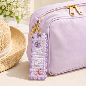 May include: A light purple rectangular pouch with gold-tone zippers and a matching scalloped fabric keychain tag. The tag features a purple gingham pattern, the name MIA embroidered in white, and small embroidered butterfly and star motifs.