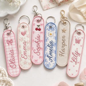 Puede incluir: Seis llaveros de tela rectangulares con cierres de mosquetón metálicos. Cada uno presenta nombres bordados: Olivia con un lazo, Emma con un corazón, Sophia con cerezas, Amelia con una margarita, Harper con una estrella y Lily con una mariposa.