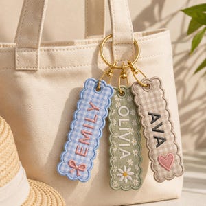 May include: Three rectangular fabric keychains with scalloped edges attached to a gold metal ring. The blue gingham keychain features the name EMILY and a bow. The green floral keychain features the name OLIVIA. The beige gingham keychain features the name AVA and a heart.