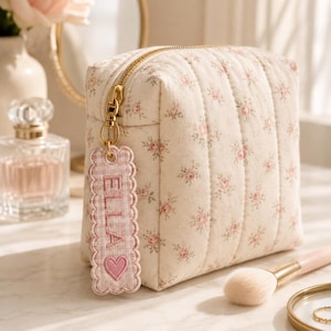 May include: A quilted cream-colored cosmetic bag featuring a small pink floral pattern and a gold zipper. Attached to the zipper pull is a scalloped, pink and white gingham fabric tag embroidered with the name ELLA and a pink heart icon.