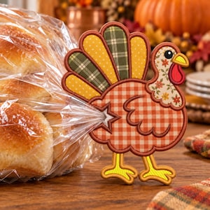 May include: A fabric turkey-shaped bread bag clip featuring a brown and white gingham body, a floral neck, and a tail made of alternating green plaid and yellow polka-dot fabric segments. The clip has yellow feet and a star-shaped opening.