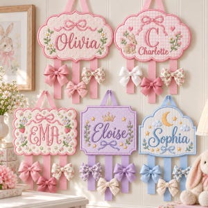 May include: Five embroidered wall hangings with scalloped edges, each featuring a name and hanging ribbons decorated with fabric bows. The designs include floral patterns, a rabbit, a crown, and a moon, rendered in pastel pink, purple, and blue.