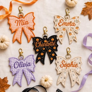 May include: Five embroidered bow keychains with gold-tone lobster clasps. The bows feature various patterns including orange gingham, pumpkin prints, black with pumpkins, purple gingham, and ghosts, each personalized with names Mia, Emma, Charlotte, Olivia, and Sophie.