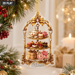 May include: A flat, bell-shaped acrylic Christmas ornament featuring a gold-toned, ornate three-tiered stand. The stand displays miniature cakes, pastries, a teapot, a teacup, and a gift box, accented with pink flowers and the word Name.