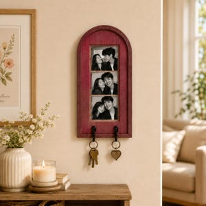 Op de afbeelding: Een verticale, boogvormige houten wandorganizer in een donkerrode afwerking met een centraal rechthoekig frame dat drie gestapelde zwart-witfoto's van een koppel bevat. Aan de onderkant zijn twee zwarte metalen haken bevestigd voor sleutels.