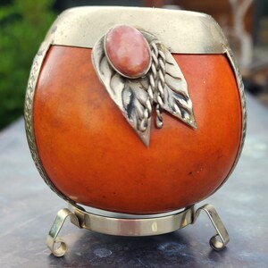 May include: A round orange mate gourd featuring a silver-toned metal rim and base. The gourd is decorated with a silver-toned metal leaf ornament and a smooth, oval-shaped pink stone set at the top. The base has three curved metal legs.