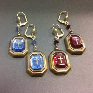 May include: Three pairs of gold tone earrings with a cross design. The earrings feature a blue or red crystal with a gold cross in the center. The earrings are on a gray background.