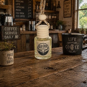 May include: A small clear glass bottle with a light-colored wooden cap and a hanging rope, featuring a label that reads Red Dirt Scents with a bull skull graphic. The bottle contains a pale liquid and is suspended by a braided cord.