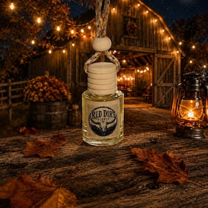 May include: A small clear glass bottle with a light-colored wooden cap and a braided hanging cord. The label features a black circular graphic with a skull and the text Red Dirt Scents. The bottle contains a pale yellow liquid.