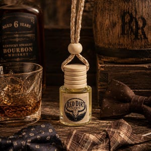 May include: A small glass bottle of fragrance with a light wooden cap and a beige rope hanger. The label features the text Red Dirt Scents with a bull skull graphic. The bottle contains a pale yellow liquid and is suspended by a wooden bead.