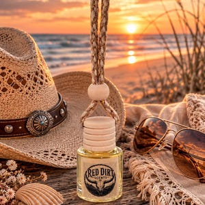 May include: A small glass car air freshener bottle with a wooden cap and a hanging rope. The label features a bull skull graphic and the text Red Dirt Scents. The bottle is positioned with a straw hat, sunglasses, a seashell, and a starfish.