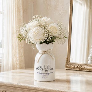 May include: A white ceramic vase with a scalloped rim and a cream ribbon tied around the neck. The vase features a black graphic of mountains, a cross, and the text Faith Can Move Mountains Matthew 17:20 and the name Olande, filled with white flowers.