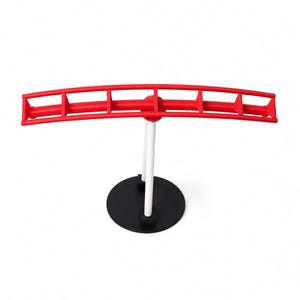 May include: A red curved horizontal bar with vertical dividers supported by two white vertical rods attached to a circular black base. The structure features a segmented design with a hollow frame, providing a stable stand for display purposes.
