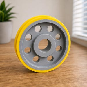 Puede incluir: Un componente de rueda circular con un borde exterior amarillo brillante y un disco interior gris mate. La sección central gris contiene un orificio central grande rodeado por ocho recortes circulares más pequeños dispuestos en círculo.