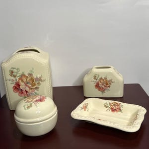May include: A four-piece cream-colored ceramic bathroom set featuring a floral pattern with orange, pink, and green tones. The set includes a tall tissue box cover, a smaller rectangular tissue box cover, a round lidded jar, and a soap dish.