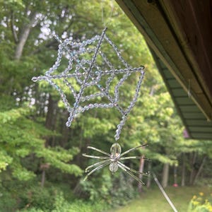 May include: A hanging ornament featuring a spider web crafted from clear, iridescent faceted beads connected by thin wire. A matching beaded spider with a light yellow oval body and clear bead legs hangs from the centre of the web structure.