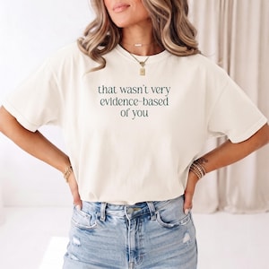 Op de afbeelding: Een crèmekleurig t-shirt met korte mouwen en de donkergroene tekst, that wasn't very evidence-based of you, gedrukt in een gecentreerd schreeflettertype op de borst van het kledingstuk, gecombineerd met een lichtblauwe spijkerbroek.