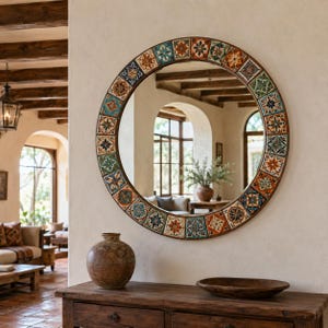 May include: A round wall mirror featuring a decorative frame composed of square tiles with intricate floral and geometric patterns in shades of blue, orange, green, and white. The mirror reflects a room with arched windows and wooden beams.