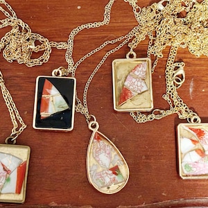 May include: Gold-toned necklaces with rectangular and teardrop pendants. The pendants feature colorful, patterned ceramic pieces in red, white, and black, set in gold frames. The necklaces rest on a wooden surface.