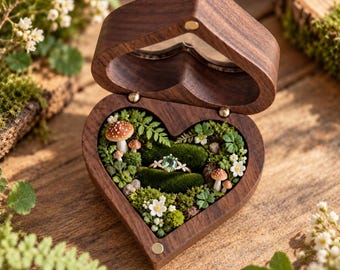 May include: A heart-shaped wooden ring box with a hinged lid, featuring a dark brown wood grain. The interior is lined with green moss, small white flowers, and miniature brown mushrooms, holding a gold ring with a green gemstone.