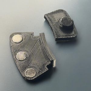May include: Two black 3D-printed components with a textured, layered finish. One piece features three circular metallic magnets embedded in a curved edge, while the other piece has a single raised circular button-like protrusion on its surface.