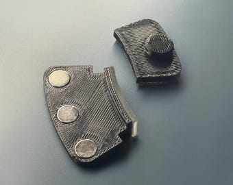 May include: Two black 3D-printed components with a textured, layered finish. One piece features three circular metallic magnets embedded in a curved edge, while the other piece has a single raised circular button-like protrusion on its surface.