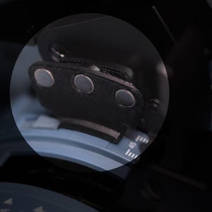 May include: A black, textured, semi-circular component featuring three embedded circular silver magnets. This piece is positioned over a blue, curved mechanical dial with white numerical markings, including the number 45, visible on the surface.